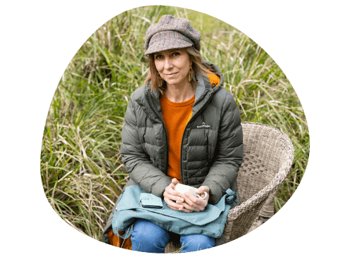 Tanya Hollis sits in a cane chair in an outdoor bush setting. She is holding a cup of coffee and is wearing a tweed cap and puffer jacket.